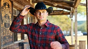 Young cowboy tipping his hat in a rustic western setting with wooden buildings in the background
