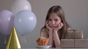 Upset little girl looking sad because celebrating birthday alone over background of gifts and balloons. Lonely holiday with no friends during pandemic and lockdown coronavirus covid-19, new normality