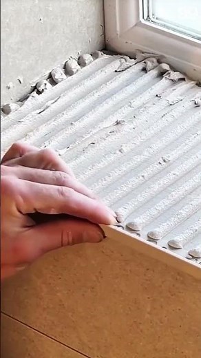 Notched trowel spreads adhesive near open window frame
