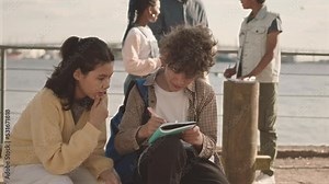 Medium slowmo of multiethnic classmates chatting while doing school project on alternative energies outdoors at sunny quay with their teacher standing with windmill model in background