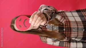 Vertical footage of serious young brunette woman 25s holding unsatisfied sad face emoticon, posing isolated over plain red color background wall in studio. Concept angry, stressed, negative emotion