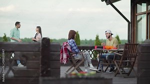 Two young couples talking on country house terrace after dinner. Emotional man telling story to girlfriend sitting at table. Cheerful couple talking over beverages leaning on railing in background