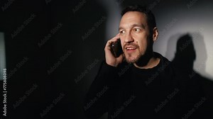 A man with facial hair is chuckling on the phone in a dimly lit room
