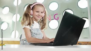 Little young cute girl using laptop to working or playing games. Student working on computer while looking and smiling at camera. Caucasian child attend online class by using headphone. Erudition.