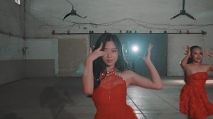 Three women in red dresses performing a dance routine in an industrial space. The central dancer poses gracefully, while the others mirror her movements. Soft lighting enhances the scene.