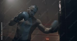 Brutal shirtless young and angry Caucasian strong male boxer punching bag in gym at ring and training. Man boxing and exercising concept.
