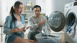 Beautiful Young Couple Sit Next to a Washing Machine at Home. They Laugh and Have a Friendly Argument While Loading the Washer with Dirty Laundry. Bright and Spacious Living Room with Modern Interior.