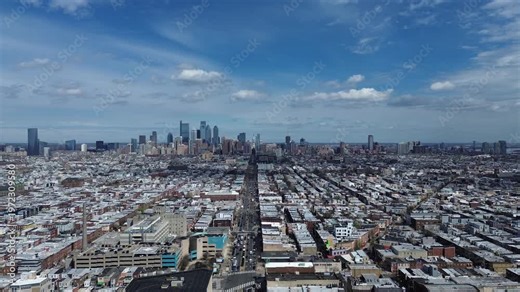 Skyline axis corridor along S Broad St frames dense South Philly blocks converging toward downtown high rise core. Flat roofs, brick gradients, bright directional light show structure, Philly, PA