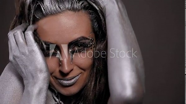 A gorgeous female model with silver body art on her body, bright makeup and silver hair poses for the camera, a chain hanging around her neck. Fashion.