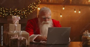 Real Santa Claus using new technology for communication with children, receiving mail or wish list. Cheerful working on laptop and smiling while sitting at his chair with fireplace and Christmas Tree