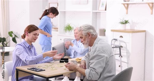 Woman playing dominoes with senior man at table and care worker helping her patient at retirement home, selective focus