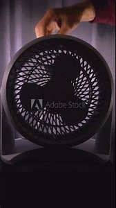 Vertical video social media format – Closeup of a man’s hand turning on a small portable plastic cooling fan, with a blade propeller, in a darkened room with closed curtains.