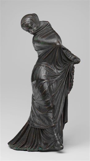 At least 2,200 years old, this bronze statuette of a veiled and masked dancer is attributed to the Hellenistic period of Ancient Greece, and is said to have come from Alexandria. The figure is shown in complex motion, her undergarment falling in heavy folds beneath a lightweight mantle drawn taut over her head and body. A sheer veil covers her face, visible at the edges and cutouts for the eyes, while her extended foot reveals a laced slipper. The interaction of layered garments conveys both mov