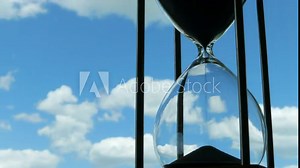 Hourglass Clouds Passing of Time Lapse. An hourglass in front of a bright blue sky with puffy white clouds passing. Shot in timelapse, passage of time concept.