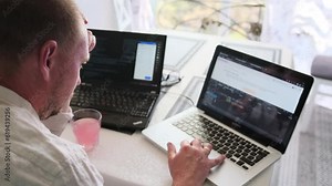 Focused man working on two laptops typing program code on keyboard during the daytime, side back view. IT specialist, programmer work in home office. Freelancer and work at home concept.