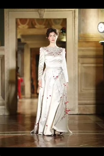 Un capolavoro di forza e bellezza: l'abito creato da Veronica per la sfilata contro la violenza sulle donne. Realizzato in raso prezioso color argento, con maniche lunghe e silhouette fluida che evoca grazia e resilienza. Il vero cuore? I ricami interamente fatti a mano: delicati fili d'argento intrecciati a gocce e petali rossi che scendono come lacrime di sangue trasformate in arte, simbolo di lotta, rinascita e empowerment femminile. Ogni punto è un atto d'amore, cucito con passione da mani a