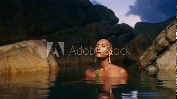 LGBTQIA black person in luxury dress dives out of water in natural ocean backwater pool. Wet gender fluid ethnic stylish model wears jewellery dressed in open outfit in tropical rocky lake at night.