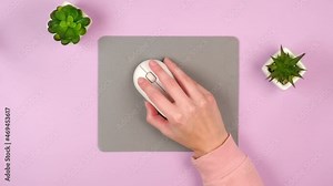 A female hand controls a white computer mouse clicks on buttons and scrolls on a gray jacket on a pink background with green plants home office