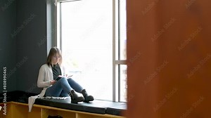student seated by large window in university library immersed in reading soft natural light quiet atmosphere provide perfect setting for her focused study with touch of warmth from colorful interior
