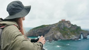 Traveler woman is making film photo to retro camera and observing beautiful view to cliffs and Atlantic ocean during holidays or vacations traveling and exploring and discover Nature world and Basque