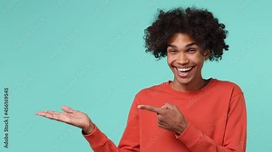 Promoter smiling happy fun young curly african american man 20s wears azure t-shirt pointing on empty palm with copy space place mock up isolated on plain pastel light blue background studio portrait