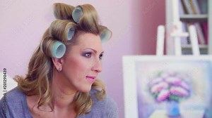 Close up portrait of attractive model with curlers posing in the room. She is just looking and smiling. Then she taking off shirt from her shoulder. Then photographer comes to her.