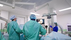Rear view of two surgeons standing at operational table. Doctors use technological devices at control the surgery process on the screen.
