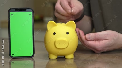 yellow piggy bank smartphone green screen hands inserting coin, close-up fintech concept showing freelancer using app to save, contrast between analog coin jar and digital interface on bright