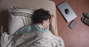 A young adult man sleeps face down on a mattress on the floor next to his laptop and smartphone. View from above
