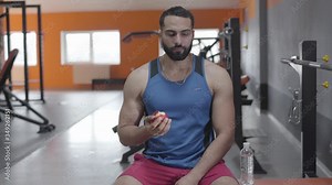 Middle shot of muscular Middle Eastern man eating apple in gym. Portrait of handsome young sportsman having break in training. Lifestyle, confidence, healthy eating, sport.