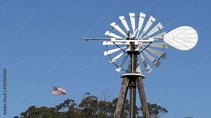 Classic retro windmill, bladed rotor and USA flag against blue sky. Vintage water pump wind turbine, power generator on livestock ranch or agricultural farm. Rural symbol of wild west, rustic suburb.
