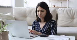 Concentrated young mixed race asian woman involved in financial calculations, paying utility bills in computer online banking application, managing monthly budget or summarizing household expenses.