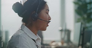 Modern call center office: Side view of Asian technical support specialist talking with customer using headset. Female hotline operator uses computer providing online assistance to client. Portrait.