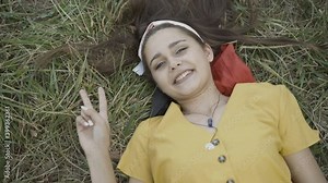 Beautiful young hippie woman lying on green meadow showing peace gesture. Portrait of relaxed carefree Caucasian lady looking at camera and smiling. Counterculture of 1960s.