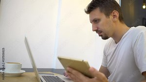 4K Caucasian man is busy to using laptop and tablet for work in a coffeeshop. Surfing the internet and touch screen to chat and contact people. Checks social network and emails on his computer-Dan