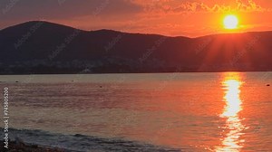 The sun is setting over the ocean, casting a warm orange glow on the water with mountains in the background creating a breathtaking natural landscape