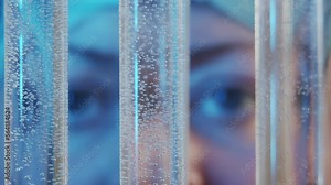 Slow motion focused eyes watching the diffusion of liquids in test tubes in the foreground in blur. A woman works in a laboratory. Rolling focus from test tubes to eyes closeup.