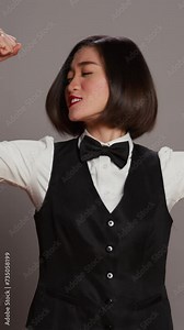Vertical Video Asian hotel concierge flexing arms muscles in grey studio, showing off her strength and power in front of camera. Receptionist with bow and uniform standing with biceps flexed. Camera B