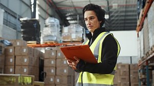 Woman in a reflective jacket taking stock in a fulfilment warehouse, she looks around assessing the inventory while reading a file. Inventory control is an essential part of her job as a warehouse man