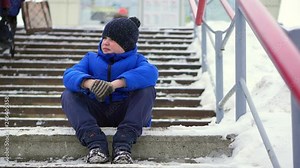 Boy teenager in blue down jacket lost in the city. He sits on a cold staircase, looks around.
