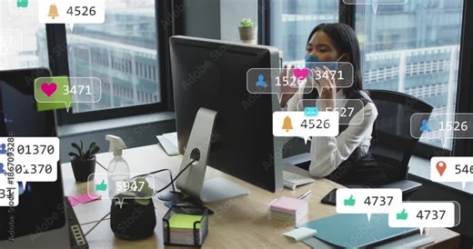 Animation of social media icons over asian woman wearing face mask using computer at office