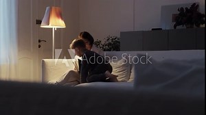 Grandmother and grandson are sitting on a white sofa and reading a book together. A cozy evening at Grandma's. Senior lady and her grandson spend time together.