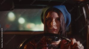 Beautiful female driver lighting cigarette and smoking behind steering wheel during night ride in vintage car with blurred city lights in background