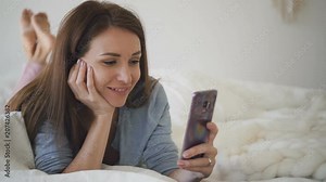 Happy woman talking, saying goodbye looking at smartphone screen lying on bed, beautiful young female is communicating in online messenger using phone, having time in bedroom. Concept: video calling