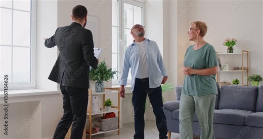 Realtor leads elderly clients on an apartment tour, discussing contract terms, purchase details in a bright living room. Real estate agent guides senior couple through apartment viewing.
