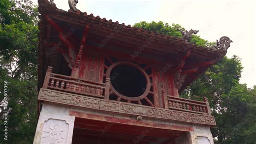 Low angle of Khue Van Cac Pavilion at the Temple of Literature in Hanoi. Iconic red wooden architecture with circular window surrounded by green trees in a historic Vietnamese site.