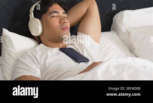 Camera tightening, framing Asian man in bed, wearing headset, phone on chest, listening, relaxing. Bed, pillows, bedding, headboard, lifestyle, minimal, stock Stock Video Footage - Alamy