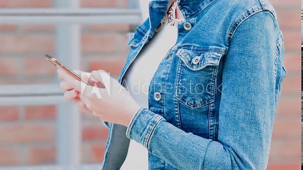 This high-quality slow-motion footage captures a detailed close-up of a young woman's hands as she interacts with her smartphone. Set against a soft-focused urban backdrop, the clip highlights modern