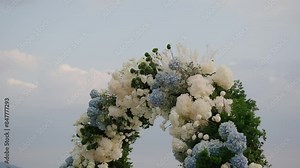 Close-up of wedding arch decorated with flowers in white and blue colors, sun rays shine between the flowers slow motion, outside wedding ceremony. wedding arch. against a cloudy
