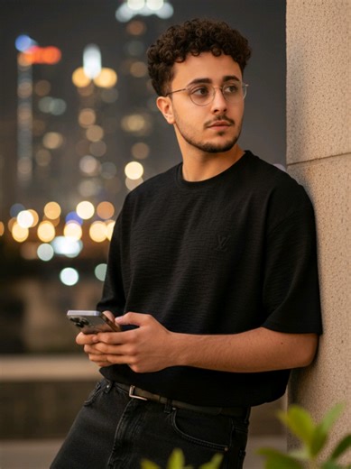 #CapCut #محتوى - من اذكا - الاصطناعي #مالي خالق-حط-الهاشتاج الشعب - الصيني - ماله - حل 🤣🤣🤣🤣‏@إسحاق عبدالله الجحدري:👇🏻👀👀👀👀👀👀👀👀👀👀👀👀 : Ultra-realistic 8K portrait of a handsome slim-fit skinny young boy (face reference from image) with curly hair. Wearing a black texture oversized LV tshirt, black loose jeans. Looking thoughtfully off-camera, holding a iphone 17 Pro, leaning on a wall. Blurred green leaves in foreground. Flawless glowing skin, vibrant bokeh city lights background,